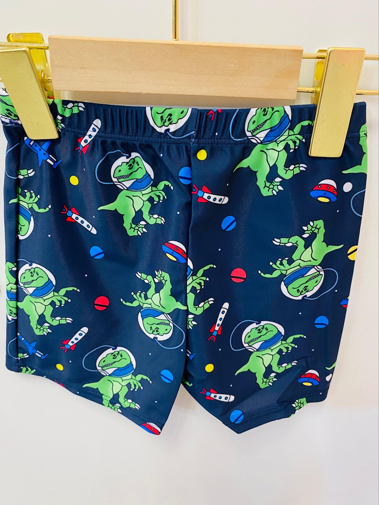 Navy swim shorts with green dinosaur astronaut design on a wooden hanger.