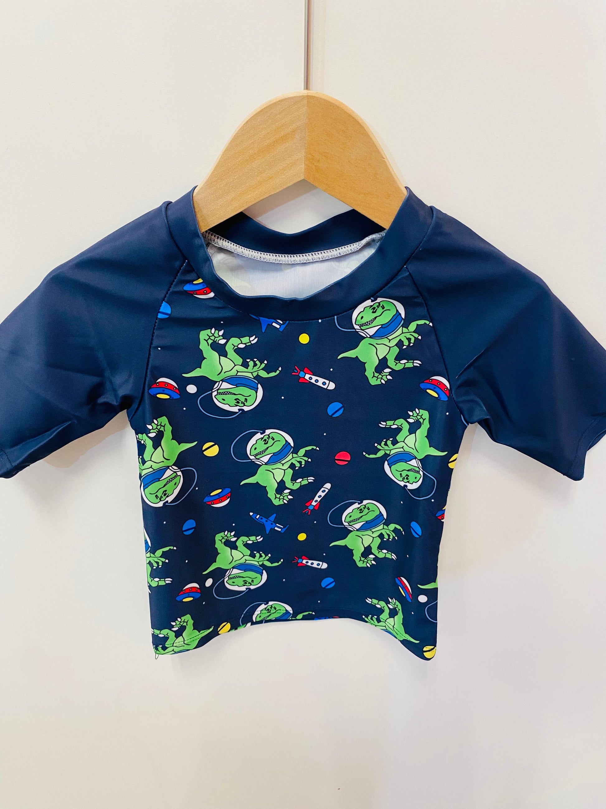 Navy blue baby shirt with green dinosaur print on a wooden hanger against a white background